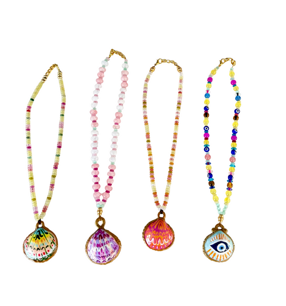 Four colorful beaded necklaces with shell-shaped pendants are displayed side-by-side against a plain off-white background. Each necklace features a different vibrant design on the pendant, including a green and yellow patterned shell, a pink and purple patterned shell, a pink and orange patterned shell, and a white shell with a blue and black eye design.